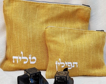 May include: Two small, black, leather tefillin bags with gold Hebrew writing. The bags are sitting on a white surface in front of two yellow fabric pouches with Hebrew writing. The larger pouch has the word "Klal" embroidered on it, and the smaller pouch has the word "Tefillin" embroidered on it.