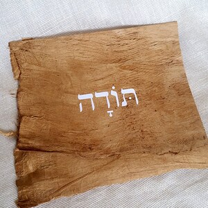 K&ouml;nnte beinhalten: Ein St&uuml;ck braunes Pergament mit dem hebr&auml;ischen Wort "תודה" in wei&szlig; geschrieben.