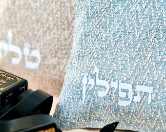May include: A light blue textured fabric pouch featuring white Hebrew text printed on the front. Beside it are black leather tefillin boxes with gold-colored Hebrew lettering and black leather straps, set against a neutral-toned fabric background.