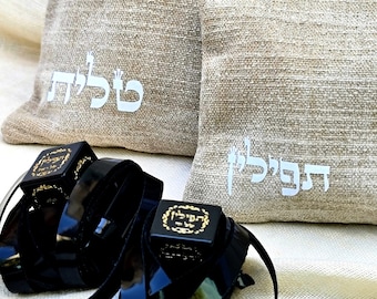 May include: Two black leather tefillin straps with gold accents and Hebrew writing on the boxes. The straps are on a beige fabric background with two beige fabric pouches. The pouches have Hebrew writing on them, "Tefillin" and "Talit".