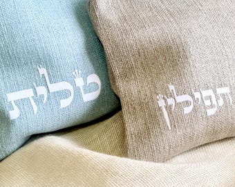 May include: Two rectangular fabric pouches, one light blue and one taupe, with white Hebrew text. The pouches are resting on a cream-colored, textured fabric. The light blue pouch is larger than the taupe one.