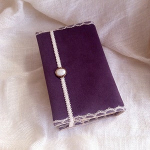May include: A purple fabric covered journal with a white lace trim and a pearl button.