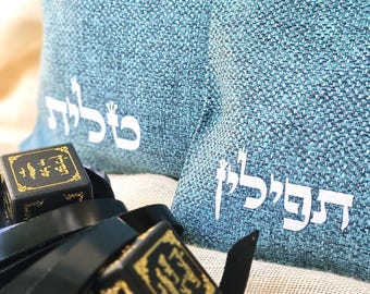 May include: A teal fabric bag featuring white Hebrew text that reads Tallit and Tefillin, positioned behind two black leather tefillin boxes with gold-colored Hebrew lettering and attached black leather straps, all set against a light beige fabric.