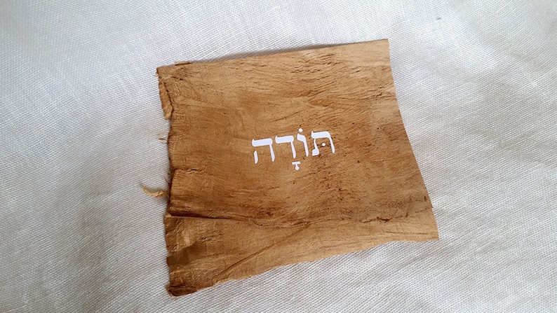 K&ouml;nnte beinhalten: Ein St&uuml;ck braunes Pergament mit dem hebr&auml;ischen Wort "תודה" in wei&szlig; geschrieben.