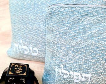 May include: Two light blue textured fabric pouches with white Hebrew text embroidered on the front, accompanied by two black rectangular boxes featuring gold decorative borders and Hebrew lettering on the top surfaces with black ribbon accents.