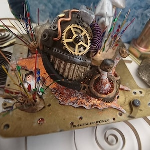 May include: A steampunk-inspired snail sculpture with gears, copper pipes, and a glittery orange base. The sculpture is surrounded by colorful wires and a white mushroom. The base is labeled "Rougegearartisan".