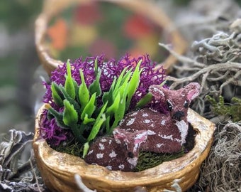 May include: A small, brown and white deer figurine is nestled in a halved walnut shell. The shell is filled with green and purple artificial plants.