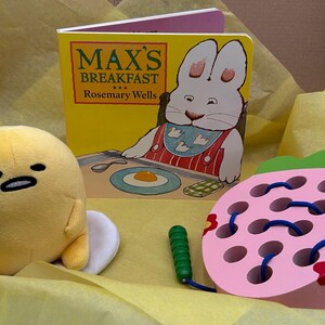 May include: A yellow plush toy shaped like an egg with a white shell, a pink wooden strawberry toy with blue string, and a children's book titled "Max's Breakfast" by Rosemary Wells.