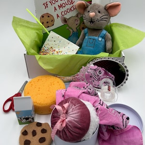 May include: A white box filled with items for a children's book-themed gift basket. The items include a plush mouse, a book titled "If You Give a Mouse a Cookie", a cookie, a milk carton, a sponge, a pink bandana, a pink velvet pouch, a small white box, a pair of scissors, and a straw.