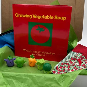 May include: A gift box containing a red book titled "Growing Vegetable Soup" with a tomato illustration, vegetable-shaped figurines, and socks with a mushroom pattern. The box is lined with green tissue paper.