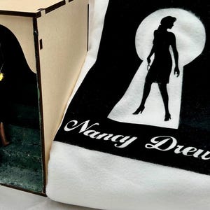 May include: A wooden box with a cutout scene of a woman in a green dress holding a lantern, climbing stairs. Next to it is a black and white graphic of a woman in a keyhole with the words "Nancy Drew".