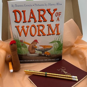 May include: A boxed set featuring a children's book titled "Diary of a Worm" with illustrations. Includes a plush worm toy with a pencil, a notebook with "Ideas" in gold lettering, and two gold pencils. The book is by Doreen Cronin.