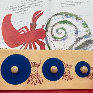May include: Wooden puzzle with three blue circles of varying sizes, each with a wooden knob. The puzzle features a red crab illustration and is set against the backdrop of an open book with a crab and seashell illustration.