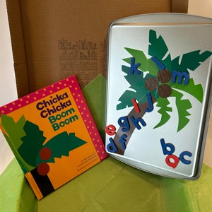May include: A colorful children's book titled "Chicka Chicka Boom Boom" by Bill Martin Jr. and John Archambault, illustrated by Lois Ehlert, is shown next to a metal baking sheet with a green palm tree design and blue and red magnetic letters.