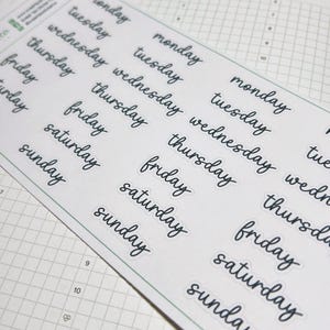 May include: A sheet of white stickers featuring the days of the week in a handwritten, cursive font. The stickers are arranged in rows and columns, with multiple instances of each day. The stickers are on top of a planner.