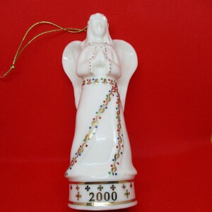 Op de afbeelding: Witte keramische engel ornament met gevouwen handen, vleugels en een gouden draad om op te hangen. De jurk van de engel is versierd met gouden, rode, blauwe en groene accenten. De basis leest "2000" in gouden letters.