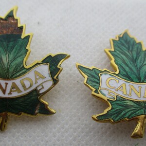 May include: Two gold-colored maple leaf pins with green enamel and a white banner that reads "CANADA".