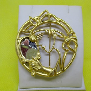 May include: A gold-toned brooch featuring a woman's profile with a mirror in her hand. The woman is depicted in a stylized art deco design.