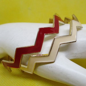 May include: Two gold-tone bangle bracelets with a zig-zag pattern. One bracelet is red and the other is cream.