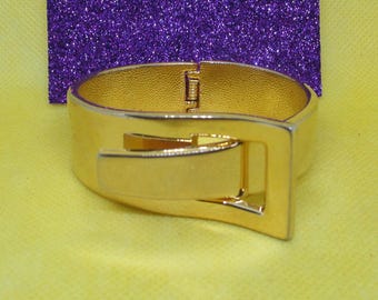 May include: A gold-toned metal bangle bracelet with a hinged clasp and a decorative buckle design.