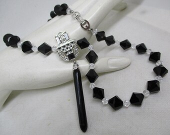 May include: A black and clear crystal necklace with a silver pendant featuring a geometric design. The pendant has a black stone hanging from it.