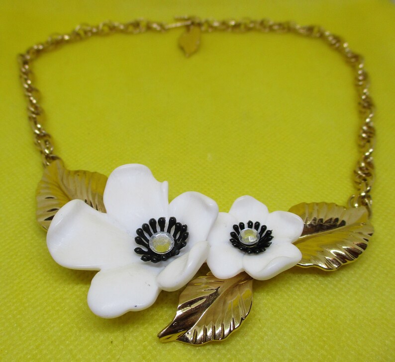 May include: A gold chain necklace with a white flower pendant. The pendant features two white flowers with black centers and gold leaves.