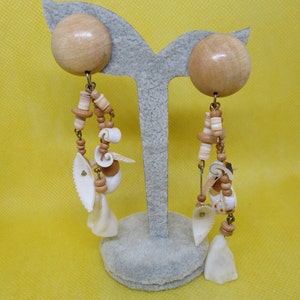 May include: Clip-on earrings with large, light-coloured wooden spheres at the top. Dangling strands below include small wooden beads, seashells, and shell-shaped pendants. The earrings have a beachy, bohemian aesthetic.