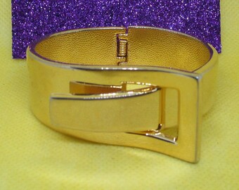 May include: A gold-toned metal bangle bracelet with a hinged clasp and a rectangular buckle design.
