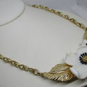 May include: A gold chain necklace with a white flower pendant. The flower has a gold center and black accents.