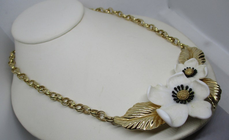 May include: A gold chain necklace with a white flower pendant. The flower has a gold center and black accents.