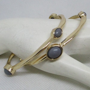 May include: Two gold bangle bracelets with gray oval-shaped stones. The bracelets are stacked on top of each other.