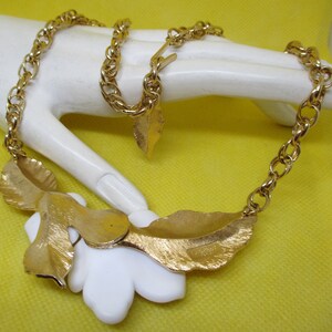 May include: A gold chain necklace with a white flower pendant and gold leaf accents. The necklace has a lobster clasp closure.