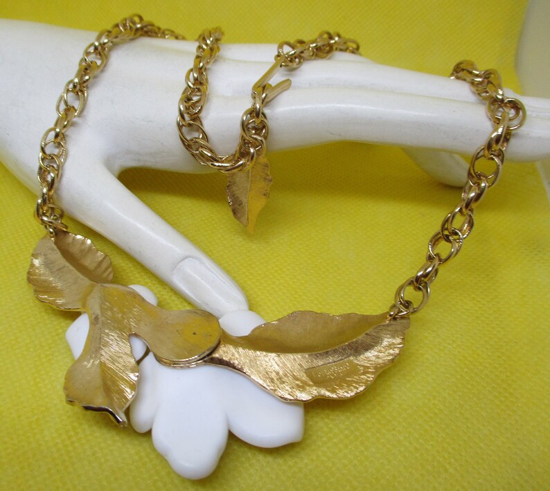 May include: A gold chain necklace with a white flower pendant and gold leaf accents. The necklace has a lobster clasp closure.