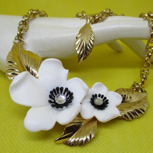 May include: A gold chain necklace with two white flower charms and gold leaf accents. The flowers have black centers with clear rhinestone accents.