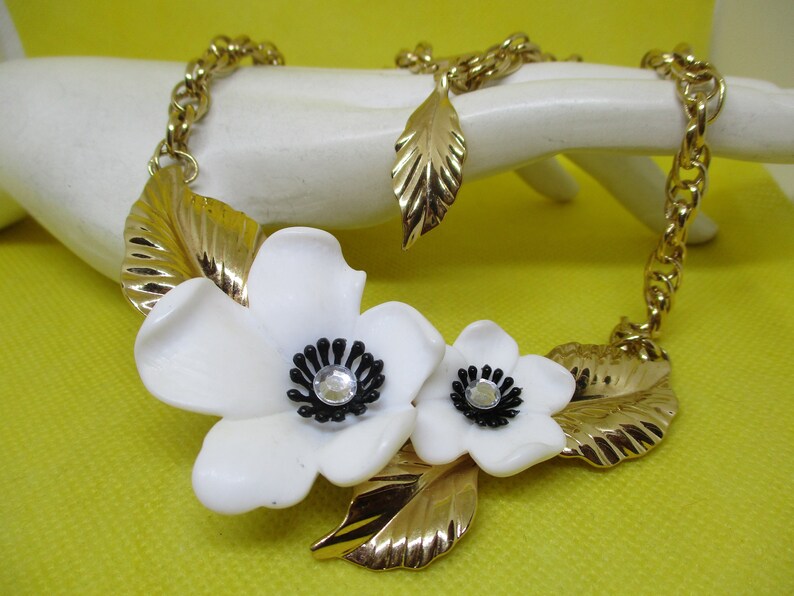 May include: A gold chain necklace with two white flower charms and gold leaf accents. The flowers have black centers with clear rhinestone accents.