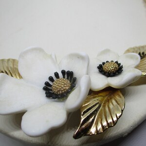 May include: A gold chain necklace with two white ceramic flowers and gold leaves. The flowers have black centers with gold accents.