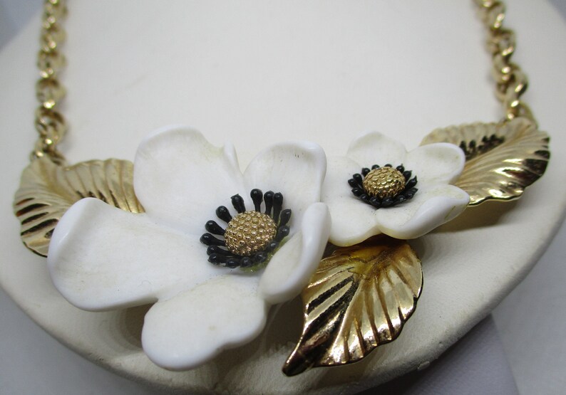 May include: A gold chain necklace with two white ceramic flowers and gold leaves. The flowers have black centers with gold accents.