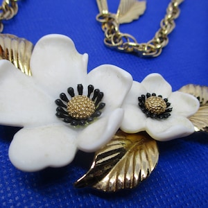 May include: A gold chain necklace with a white flower pendant. The pendant features two white flowers with black centers and gold leaves.