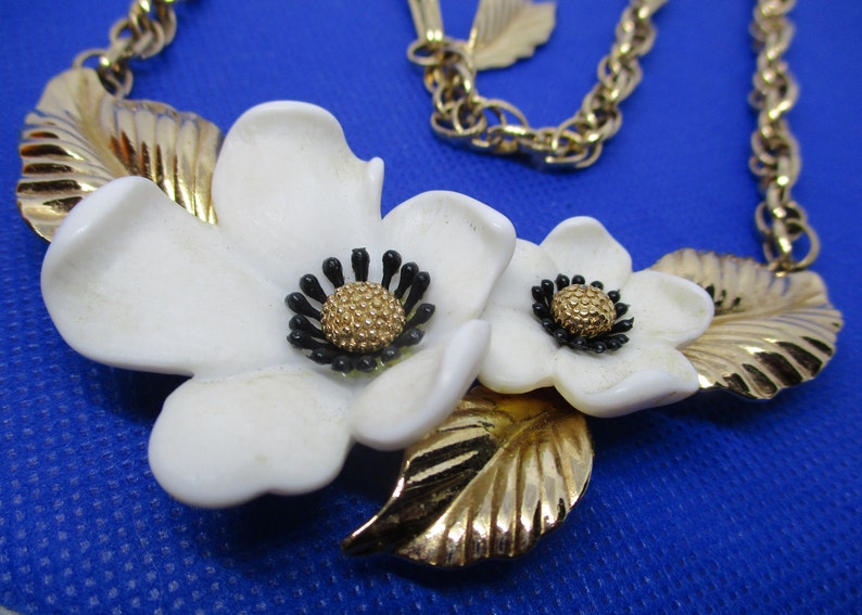 May include: A gold chain necklace with a white flower pendant. The pendant features two white flowers with black centers and gold leaves.