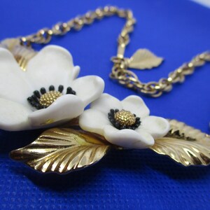 May include: A gold chain necklace with a white flower pendant. The pendant features two white flowers with black centers and gold leaves.