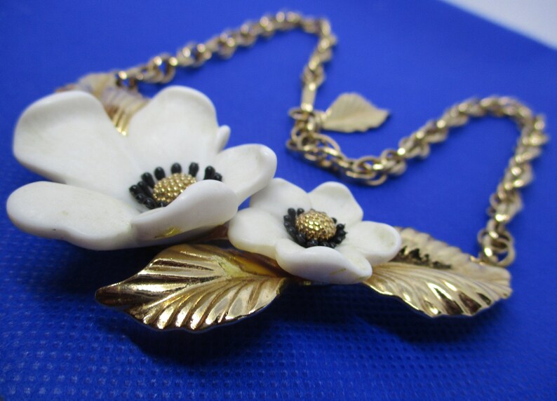 May include: A gold chain necklace with a white flower pendant. The pendant features two white flowers with black centers and gold leaves.