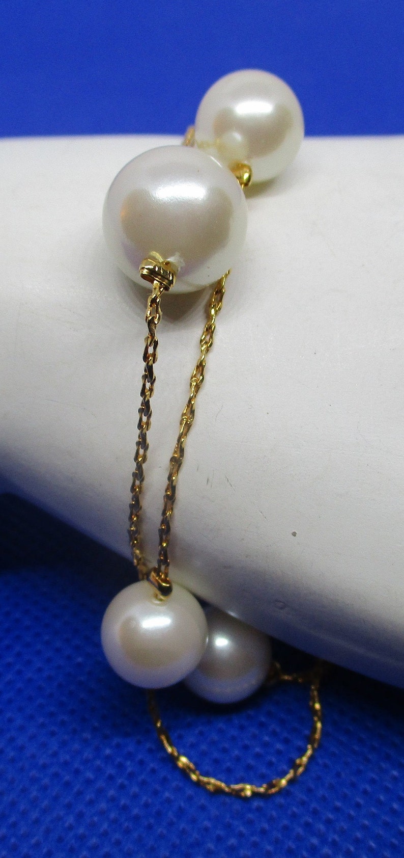 May include: A gold chain bracelet with four white pearls. The bracelet is on a white surface with a blue background.