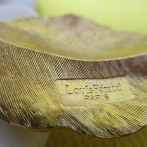 May include: Close-up of a gold-colored metal leaf-shaped object with a label that reads "Louis Feraud Paris".