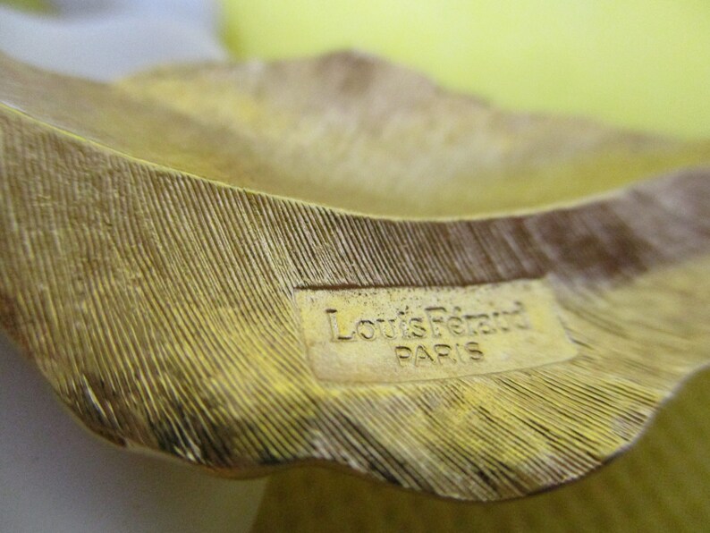 May include: Close-up of a gold-colored metal leaf-shaped object with a label that reads "Louis Feraud Paris".