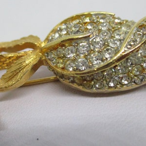 May include: Gold tone brooch featuring a single rose with a pearl accent. The rose is made of clear rhinestones and has a gold tone stem and leaves.