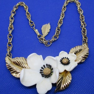 May include: A gold chain necklace with a white porcelain flower pendant. The pendant features two flowers with black centers and gold leaves.