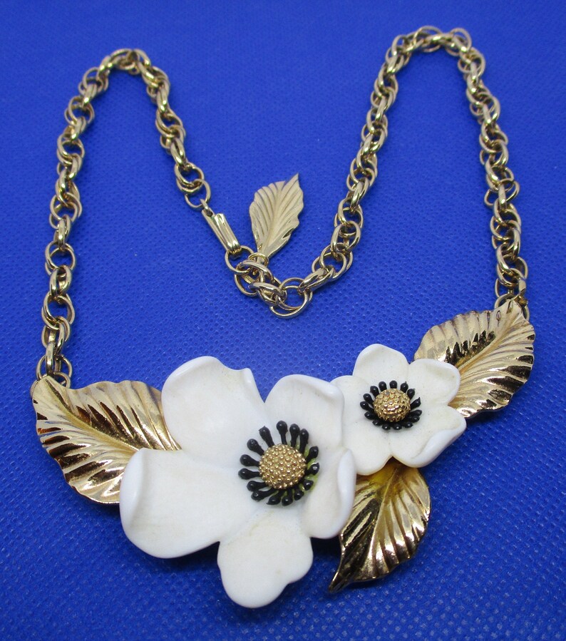 May include: A gold chain necklace with a white porcelain flower pendant. The pendant features two flowers with black centers and gold leaves.