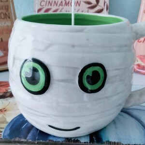 May include: A white ceramic mug with a mummy face design, featuring large green eyes and a black smile. The mug has a green interior and a lit candle. Decorative signs with text are in the background.