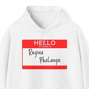 Puede incluir: Sudadera con capucha blanca con un diseño de etiqueta con el nombre en rojo. La etiqueta dice "HELLO MY NAME IS" encima del nombre "Regina Phalange" en escritura negra. La sudadera tiene un cordón y un bolsillo de canguro.