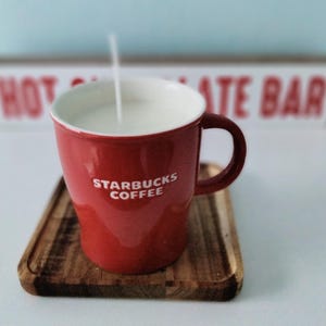 Op de afbeelding: Een rode Starbucks koffiemok met een witte binnenkant, gevuld met een witte vloeistof en een aangestoken lont. De mok staat op een kleine houten dienblad. Op de achtergrond staat een bord met de tekst "HOT CHOCOLATE BAR".
